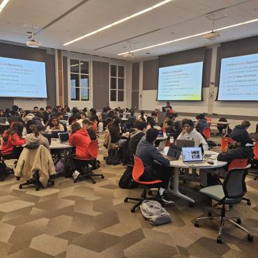 Room of students seated at circular tables with screens showing a presentation during a workshop
