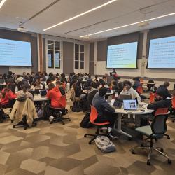 Room of students seated at circular tables with screens showing a presentation during a workshop