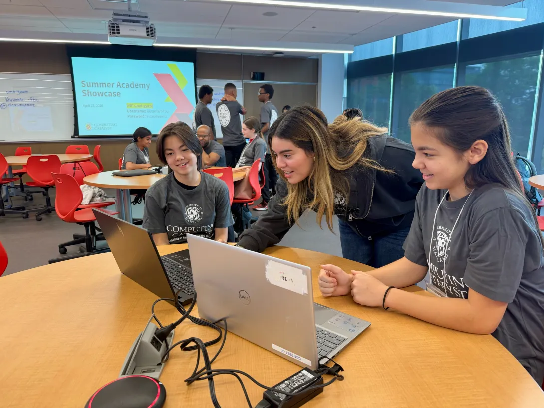 Two children working at computers with an undergraduate student looking over their shoulders