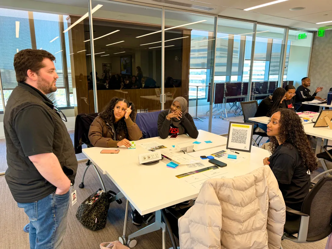 A group of RESET Scholars seated at a table while talking with an EY employee, who is standing on the left